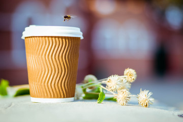 Paper cup with coffee. Dry flowers on brown background. Bright summer good morning. A bee flies near a coffee cup. Brown paper cup with plastic white lid.