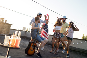 Young friends having fun at a rooftop party, playing the guitar, singing, dancing and drinking