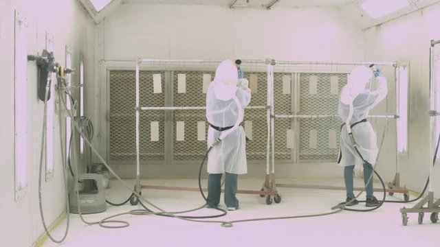 Sliding shot of 2 workers wearing respirators and coating manufactured parts inside a paint spray booth