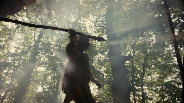 Primeval Caveman Wearing Animal Skin Holds Stone Tipped Spear Looks Around Prehistoric Forest, Ready to Hunt Animal Prey. Neanderthal Going Hunting into the Jungle. Low Angle Slow Motion Arc Shot