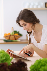 Young woman looking for a new recipe for cooking in a kitchen. Housewife is making online shopping by tablet computer and credit card