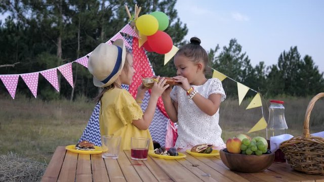cute hungry children eat baked pastries and drink it with juice at picnic in the forest during kids party on wigwam background