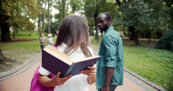 Female student in eyeglasses hit in park while walking and reading book, afro-american male apologizing