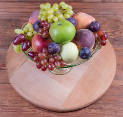 Different fruits in glass fruit vase on wooden serving board