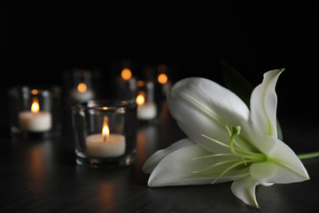 White lily and blurred burning candles on table in darkness, closeup with space for text. Funeral symbol