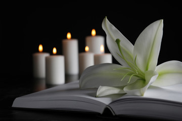 White lily, book and blurred burning candles on table in darkness, closeup with space for text. Funeral symbol