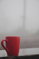 Red mug of coffee on wood table with pine tree forest in the mist and rain background.