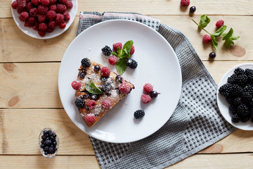 Piece of pie with blueberries, rasberry and mint for dessert on a white plate, napkin. Pieces of delicious homemade cake on a wooden boards background