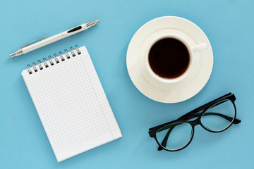 Office supplies or education concept : Top view image of notepad, glasses, pen and coffee cup on blue background. Top view with copy space.