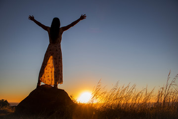 Silhouette of a beautiful woman standing on a rock with her arms spread open at sunset or sunrise.