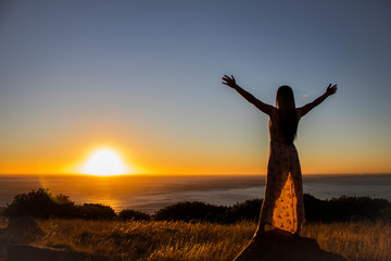 Silhouette of a beautiful woman standing on a rock with her arms spread open overlooking the ocean at sunset or sunrise.