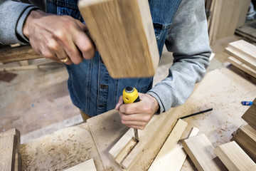 Man carpenter makes a hole in the wooden furniture detail with a hammer and chisel.