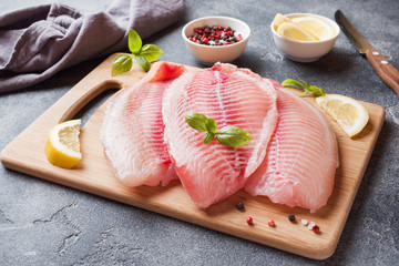 Raw fish fillet of tilapia on a cutting Board with lemon and spices. Dark table with copy space.