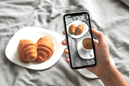 Female hand with mobile phone taking picture of tasty breakfast coffee and croissants in bed, closeup