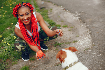 A young and stylish dark-skinned girl with red dreads walking in the summer park with dog