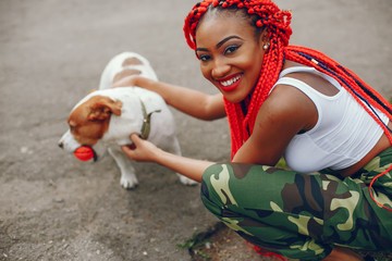 A young and stylish dark-skinned girl with red dreads walking in the summer park with dog