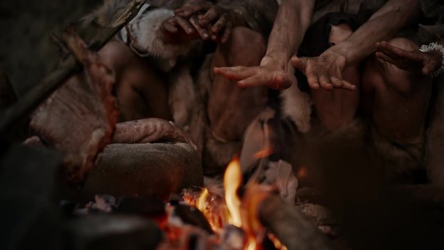 Tribe of Prehistoric Hunter-Gatherers Wearing Animal Skins Live in Cave at Night. Neanderthal or Homo Sapiens Family Trying to Get Warm at the Bonfire, Holding Hands over Fire, Cooking Food. Close-up