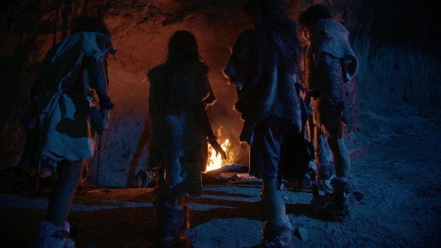 Tribe of Prehistoric Hunter-Gatherers Wearing Animal Skins Live in a Cave at Night. Neanderthal or Homo Sapiens Family Trying to Get Warm at the Bonfire, Holding Hands over Fire. Following Back View