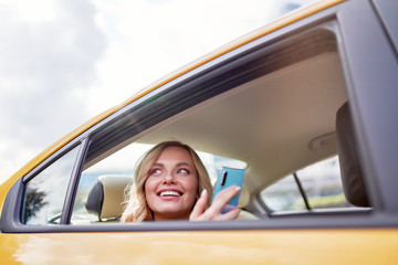 Photo of happy blonde looking at side with phone in her hand sitting in back seat in yellow taxi
