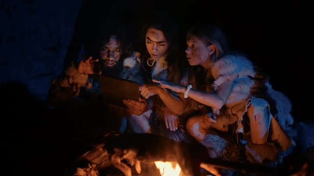 Tribe of Prehistoric, Primitive Hunter-Gatherers Wearing Animal Skins Use Digital Tablet Computer in a Cave at Night. Neanderthal or Homo Sapiens Family Browsing Internet, Watching Videos, TV Shows