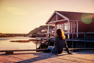 Beautiful young  girl traveler sitting on wooden pier on the background of traditional red rorbu...