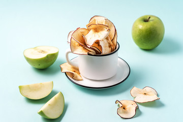 Dried apple chips in a cup and fresh apples on light blue background.