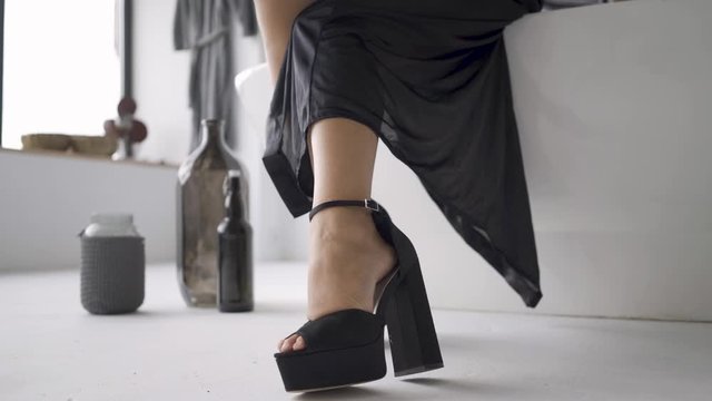 Feet of young woman in elegant black clothes with slim body sitting on teh bath in the bathroom. Bottles standing on the floor. The girl preparing for the date. Happy mood