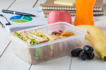 Office desk with supplies and Egg salad with sandwich slices in lunch box with fruits and orange juice.
