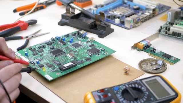 Service center employee with multimeter or multitester (also known as a VOM - volt ohm milliammeter) checks for serviceability green motherboard. Repair Shop and Electronic Devices Concept