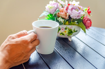 A man's hand holds a white Cup of coffee on a wooden table close-up, next to a bouquet of flowers
