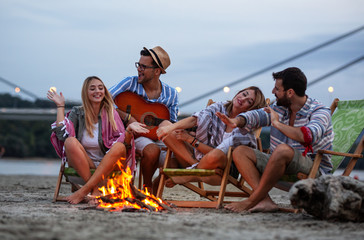 Group of friends sitting around camp fire at the beach at the evening.They play guitar and singing.