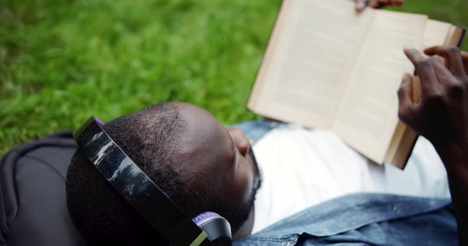 Male afro-american student in headphones lying on grass in park and reading book, literature