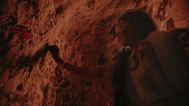 Primitive Prehistoric Neanderthal Child Wearing Animal Skin Draws Animals and Abstracts on the Walls at Night. Creating First Cave Art with Petroglyphs, Rock Paintings Illuminated by Fire. Low Angle