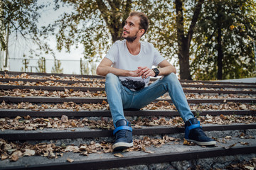 Obraz premium A young handsome man in casual clothes is sitting on the steps in the park and looking around.