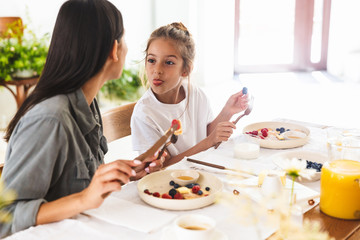 Image of amusing family mother and little daughter eating tasty pancakes together while having breakfast at home in morning