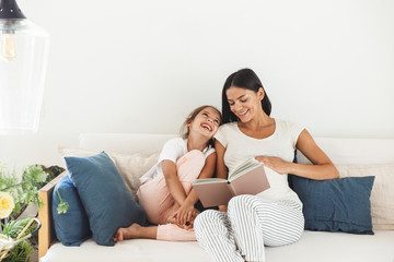 Image of joyful family pregnant woman and her little daughter reading book while sitting on sofa at...