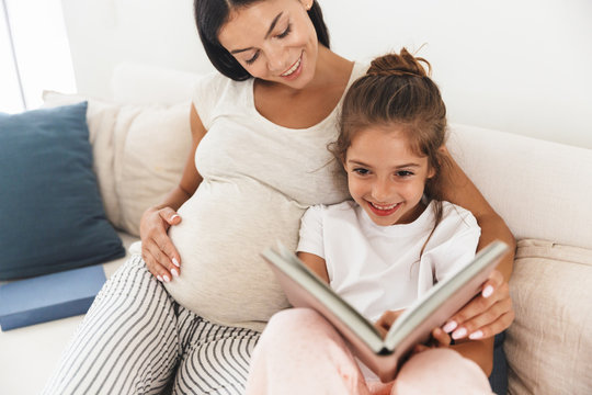 Image of cute family pregnant woman and her little daughter reading book while sitting on sofa at home