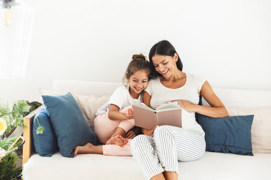 Image of attractive family pregnant woman and her little daughter reading book while sitting on sofa at home