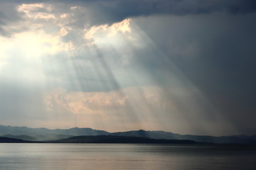 Dark sea and cloudy sky with sun rays lightning through the clouds. Green hills and mountains on the horizon. Landscape and seascape for design template, background or wallpaper.