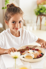 Portrait of caucasian little girl sitting at table in bright kitchen while eating pancakes with raspberry