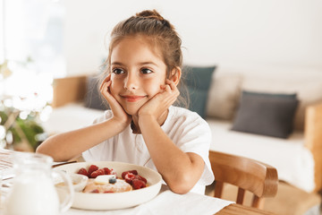 Portrait of adorable little girl sitting at table in bright kitchen while eating pancakes with raspberry