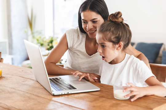 Image of pleased family woman and her little daughter smiling and using laptop computer together in apartment