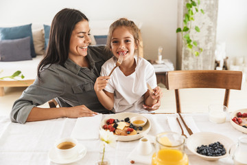 Image of joyous family mother and little daughter smiling and eating together while having breakfast at home in morning