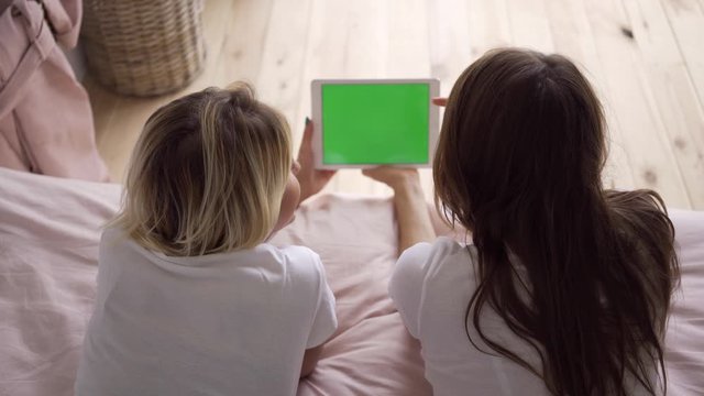 Back view of two young women in white t-shirts lying on the bed watching photos on the tablet. Leisure at home. Chromakey, green screen