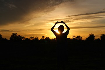 The silhouette of the boy raised his heart-shaped hands and the blue sky, golden yellow sunlight at sunset, taking a blurred photo