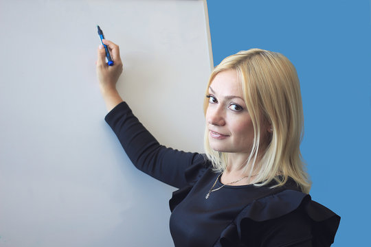 Close-up. Blond woman writes on a blackboard by a flamaster. Looks at the camera and smiles. Blue background. 