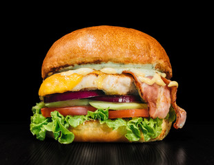 Craft burger is cooking on black background. Consist: sauce, lettuce, tomato, red onion, cucumber, cheese, bacon, air bun and breast of chicken.