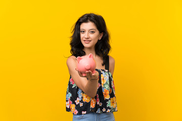 Young woman over isolated yellow background holding a big piggybank