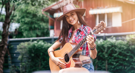 Fototapeta premium Portrait of caucasian young woman in checkered shirt and cowboy hat. She is looking at camera with a smile while playing country music using guitar outdoors. Living on a farm concept. Horizontal shot