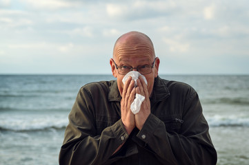 A man stands by the sea and uses a handkerchief, he has an allergy or severe flu. Concept: Runny nose or protection against infections